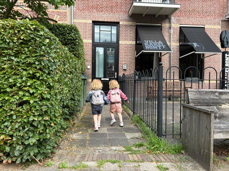 Twee jonge kinderen lopen hand in hand naar de ingang van kinderopvang Villa Kakelbont